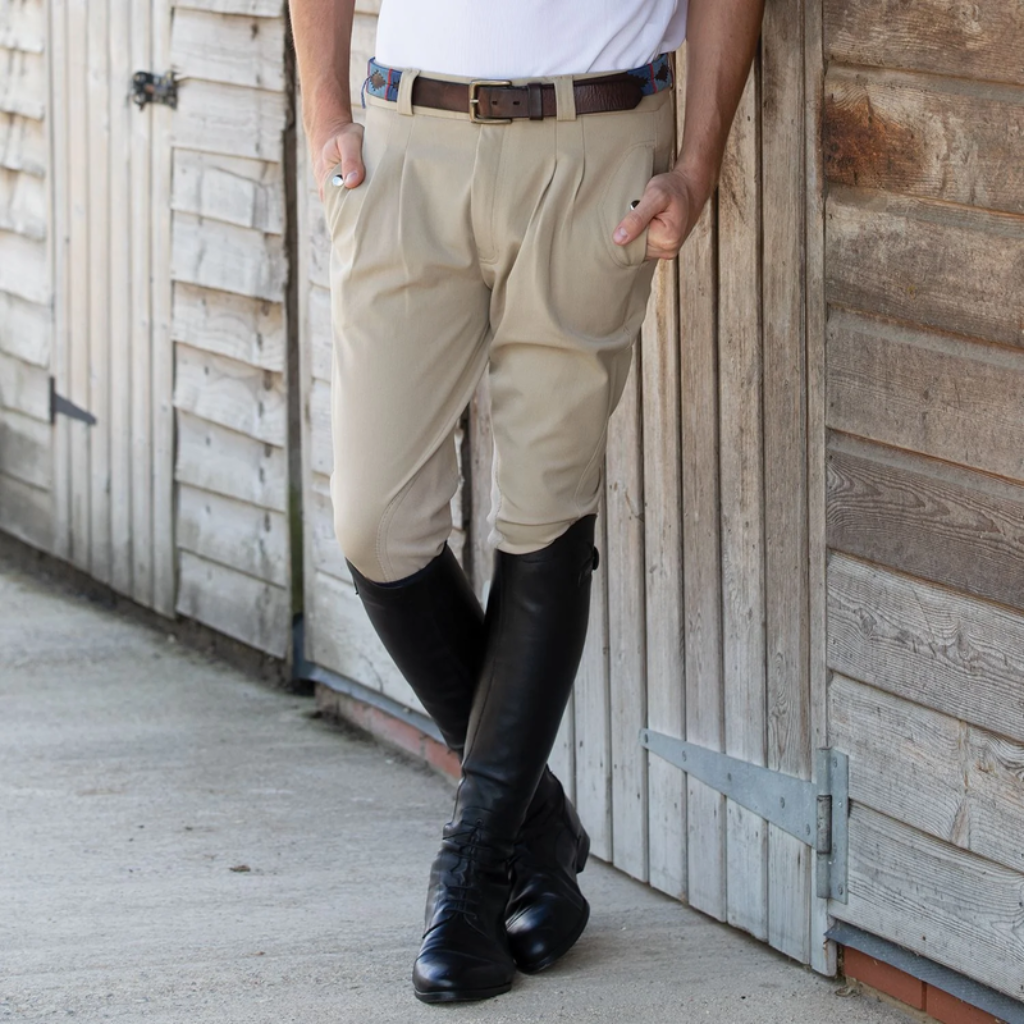 Man in tan jodhpurs and black tall boots leaning against a timber stable