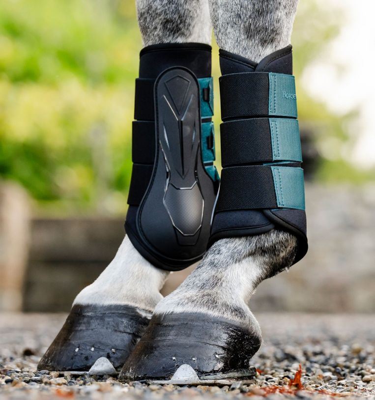 Close-up of a grey horse's front legs wearing black and teal brushing boots on a gravel yard