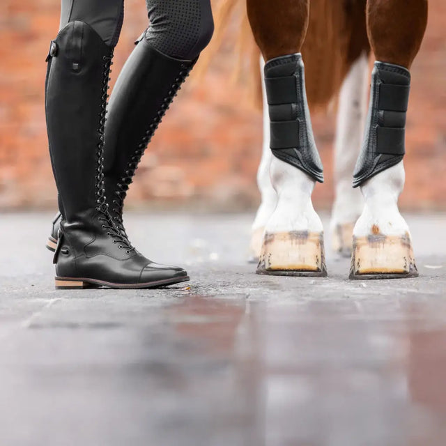 A rider in tall riding boots beside a horse in brushing boots